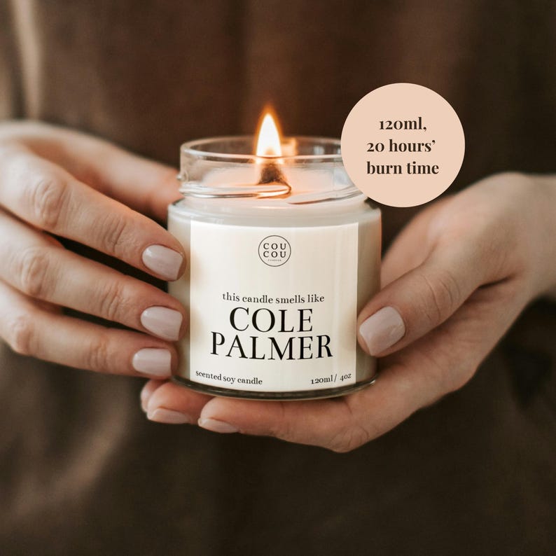 May include: A white soy candle in a glass jar with a black label that reads "this candle smells like COLE PALMER" and "120ml/4oz". The candle is lit and has a flame. A white circle with black text reads "120ml, 20 hours' burn time".