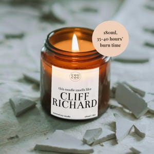 May include: A brown glass candle jar with a white label that reads "this candle smells like CLIFF RICHARD" and "Cou Cou Candles". The candle is lit and the jar is sitting on a white surface.