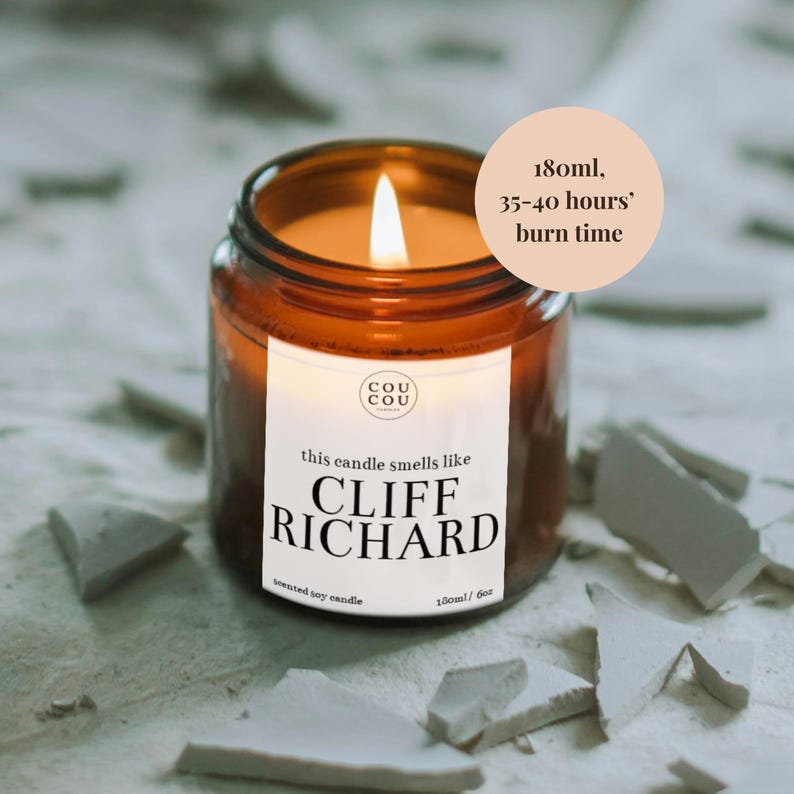 May include: A brown glass candle jar with a white label that reads "this candle smells like CLIFF RICHARD" and "Cou Cou Candles". The candle is lit and the jar is sitting on a white surface.