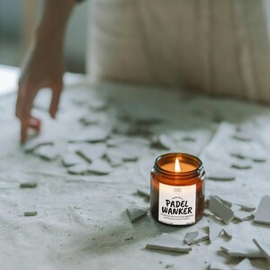 May include: A lit, amber-colored candle in a glass jar with a white label that reads "PADEL WANKER". The candle sits on a surface covered in broken pieces. The candle's flame is bright and the jar is round.
