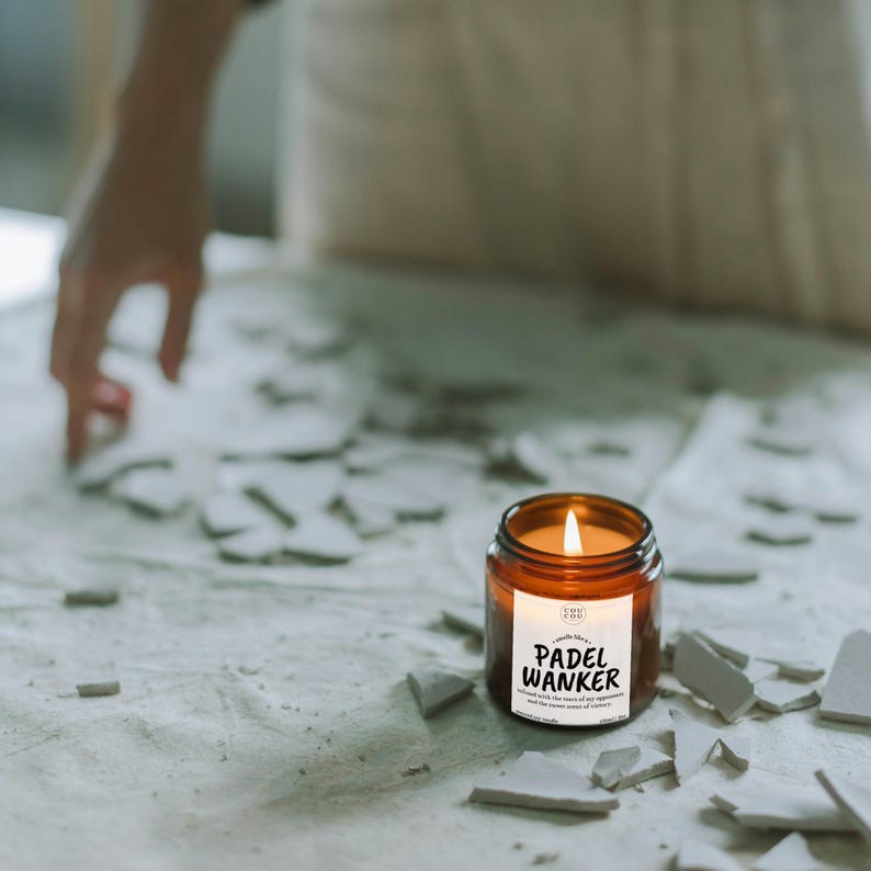 May include: A lit, amber-colored candle in a glass jar with a white label that reads "PADEL WANKER". The candle sits on a surface covered in broken pieces. The candle's flame is bright and the jar is round.
