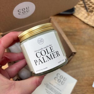 May include: A glass jar candle with a gold lid and a white label. The label says "this candle smells like COLE PALMER" and "scented soy candle 120ml/4oz".