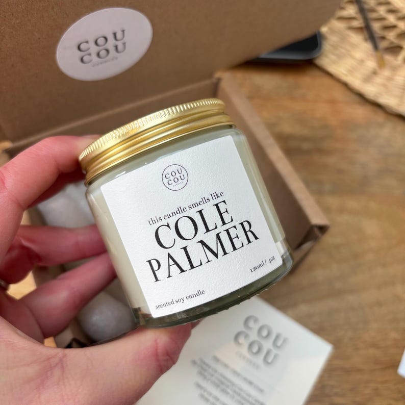 May include: A glass jar candle with a gold lid and a white label. The label says "this candle smells like COLE PALMER" and "scented soy candle 120ml/4oz".