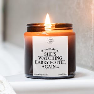 May include: A lit, scented soy candle in a brown glass jar. The label reads "smells like SHE'S WATCHING HARRY POTTER AGAIN...". The candle is 180ml and has a burning flame.