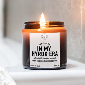May include: A lit scented soy candle in a brown glass jar. The label reads "IN MY HYROX ERA" and lists the scent as a blend of sweat, supplements, and sled pushes. The candle is 180ml / 6oz.