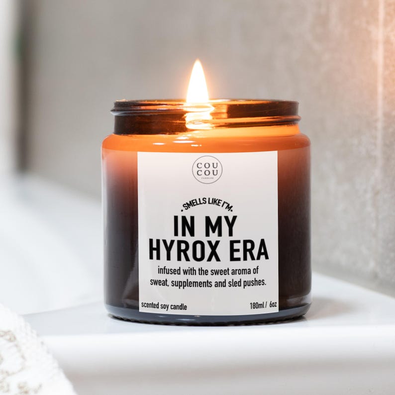 May include: A lit scented soy candle in a dark amber glass jar with a white label. The label reads: COU COU, SMELLS LIKE I'M IN MY HYROX ERA, infused with the sweet aroma of sweat, supplements and sled pushes, scented soy candle, 180ml / 6oz.