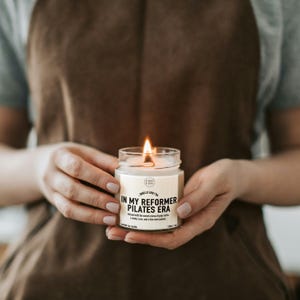 May include: A clear glass jar candle with a lit wick and a white label featuring the text "SMELLS LIKE I'M IN MY REFORMER PILATES ERA" along with "infused with the sweet aroma of grip socks, a shaky core, and a few more pulses" and "120ml / 4oz".