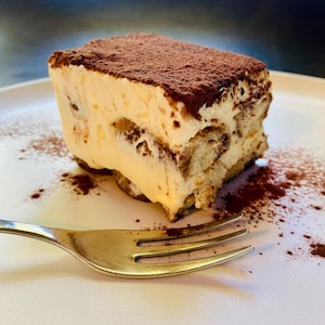 Puede incluir: Una porción de tiramisú sobre un plato blanco, espolvoreado con cacao. El postre presenta capas de crema y bizcocho de soletilla, con un tenedor plateado en el plato.