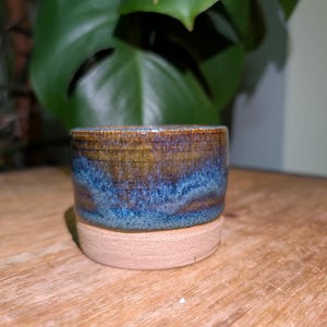 May include: A handmade ceramic pot with a blue and brown glaze, resting on a beige base. The pot is cylindrical and sits on a wooden surface. A large green plant is in the background.
