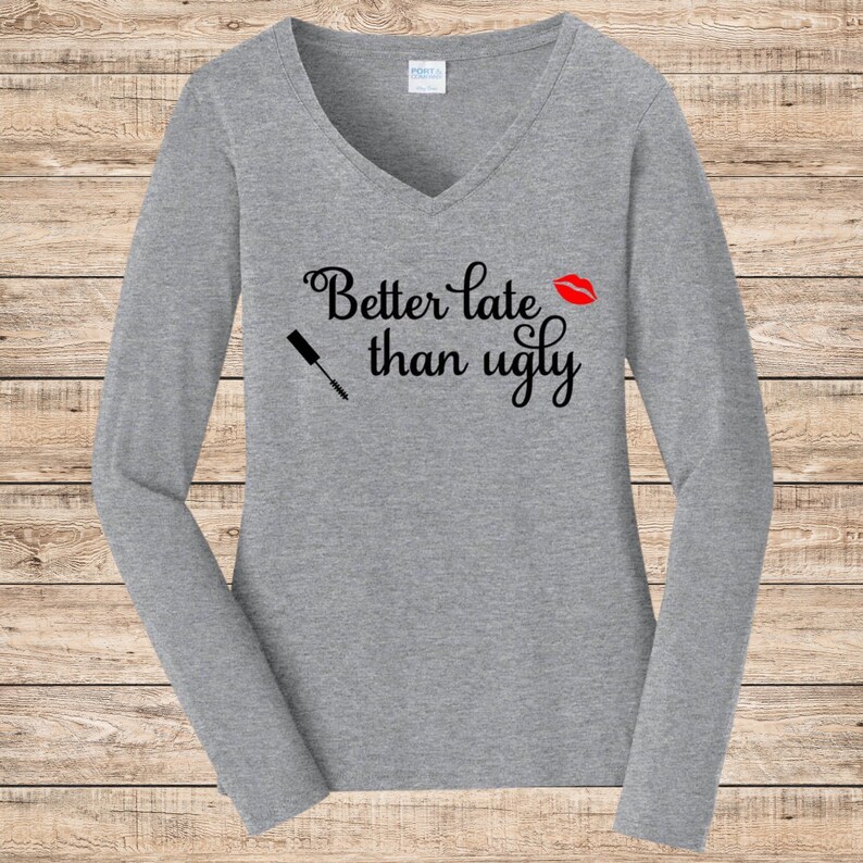 May include: A heather gray long-sleeve V-neck shirt with the phrase "Better late than ugly" in black script. A red lipstick kiss and a mascara wand are also featured on the shirt.