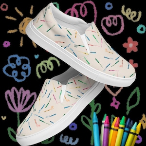 May include: Pair of cream-colored slip-on shoes with a pattern of colorful crayons. The shoes have white soles and a white elastic band across the top. The background is black with colorful crayon drawings.