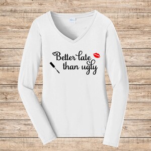 May include: White long-sleeve V-neck shirt with the phrase "Better late than ugly" in black script. A red lipstick kiss and a mascara wand graphic are also featured on the shirt.