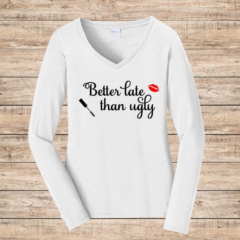 May include: White long-sleeve V-neck shirt with the phrase "Better late than ugly" in black script. A red lipstick kiss and a mascara wand graphic are also featured on the shirt.