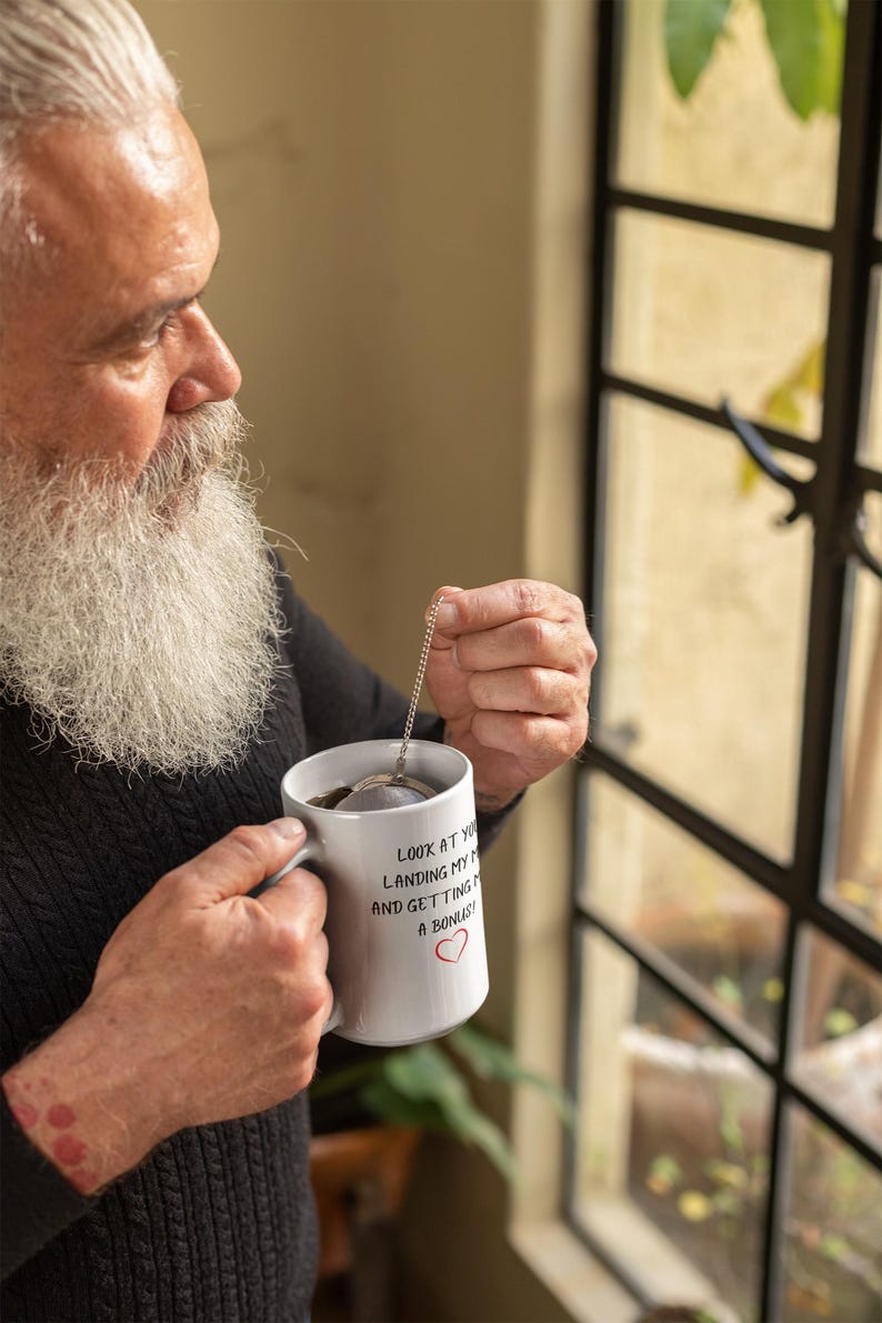 Pu&ograve; includere: Una tazza in ceramica bianca con testo nero che recita "Guardati, stai atterrando il mio aereo e mi stai facendo un bonus!" con un cuore rosso. Una persona tiene la tazza e un infusore per il t&egrave;.