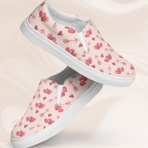 May include: A pair of white canvas slip-on shoes with a pink background and a pattern of red hearts and arrows.
