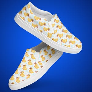 May include: A pair of white canvas slip-on shoes with a pattern of yellow rubber ducks wearing sunglasses. The ducks are surrounded by blue bubbles.