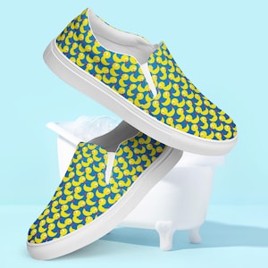 May include: A pair of white canvas slip-on shoes with a blue background and a yellow rubber ducky pattern.