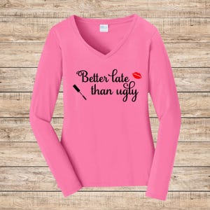 May include: A long-sleeved, pink V-neck shirt. The shirt features the phrase "Better late than ugly" in black script, along with a red lipstick kiss and a mascara wand graphic.