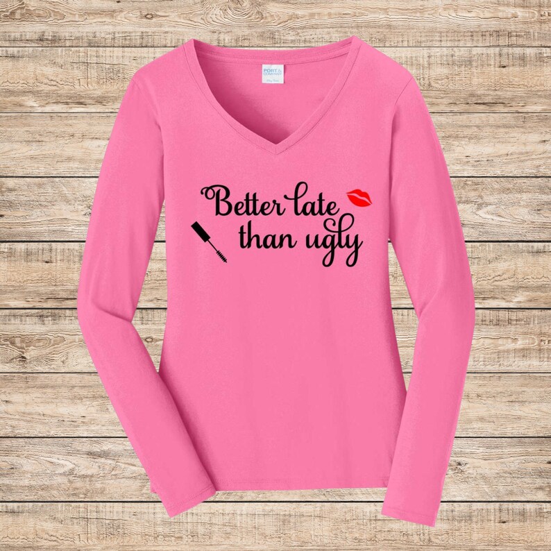 May include: A long-sleeved, pink V-neck shirt. The shirt features the phrase "Better late than ugly" in black script, along with a red lipstick kiss and a mascara wand graphic.