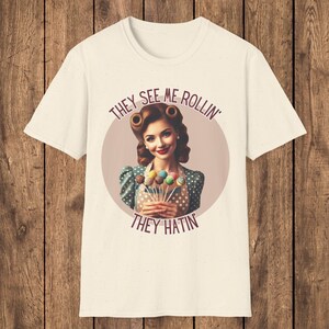 May include: White t-shirt with a retro-style illustration of a woman holding colorful cake pops. The text "They See Me Rollin' They Hatin'" is printed on the shirt.