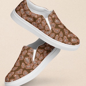 Puede incluir: Un par de zapatos de lona blancos con un estampado de esqueleto de dinosaurio marrón. Los zapatos están sobre un fondo marrón claro.