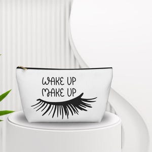 Könnte beinhalten: Eine weiße Kosmetiktasche mit dem schwarzen Schriftzug WAKE UP MAKE UP und einer grafischen Darstellung langer Wimpern. Die Tasche hat einen schwarzen Reißverschluss an der Oberkante und eine konische Form mit breitem Boden und schmaler Öffnung.