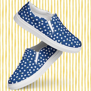 May include: Blue slip-on shoes with a white tooth pattern and white soles. The shoes have a casual design and are suitable for everyday wear. The background is a yellow and white striped pattern.