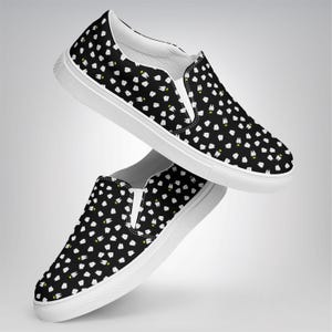 May include: Black slip-on shoes with a white sole and a pattern of white teeth with yellow accents. The shoes have a white elastic band on the top and a white interior.