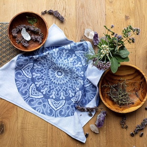 Op de afbeelding: Een witte katoenen theedoek met een blauwe en witte mandala print. De handdoek ligt op een houten oppervlak met kommen, kruiden en kristallen eromheen verspreid.
