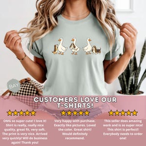 May include: Light green t-shirt featuring three cartoon ducks, each paired with a dog. The ducks are white with orange beaks and feet. The dogs are brown, black, and white. The text "CUSTOMERS LOVE OUR T-SHIRTS!" is at the bottom.