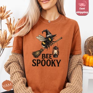 May include: Orange t-shirt with a graphic of a bee wearing a witch hat, riding a broom, and carrying a lantern. The text "BEE SPOOKY" is below the graphic. The shirt is part of a Halloween-themed collection.