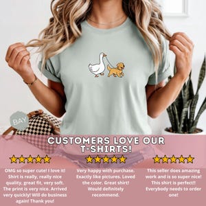 Pu&ograve; includere: T-shirt verde chiaro con un'illustrazione a cartoni animati di un'anatra bianca che porta a spasso un golden retriever al guinzaglio. Il testo "CUSTOMERS LOVE OUR T-SHIRTS!" &egrave; in basso.