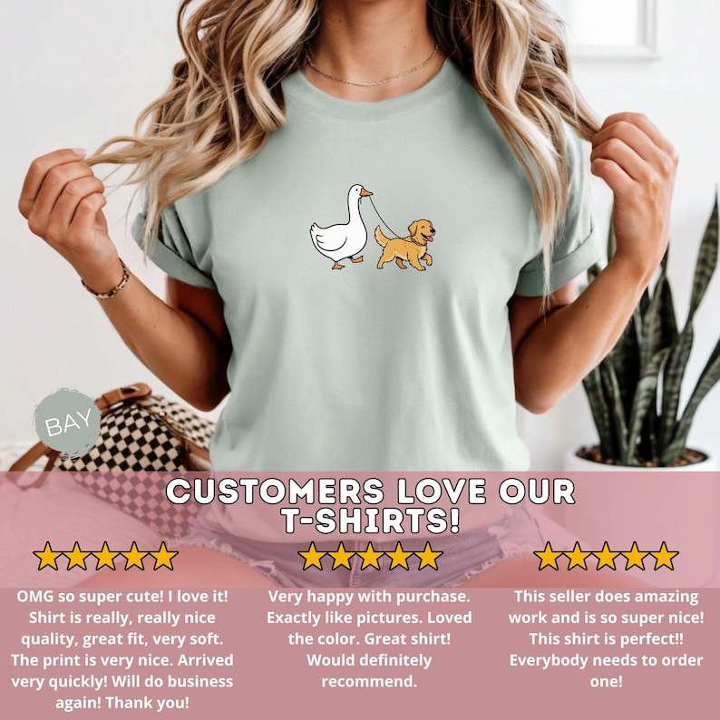 Pu&ograve; includere: T-shirt verde chiaro con un'illustrazione a cartoni animati di un'anatra bianca che porta a spasso un golden retriever al guinzaglio. Il testo "CUSTOMERS LOVE OUR T-SHIRTS!" &egrave; in basso.