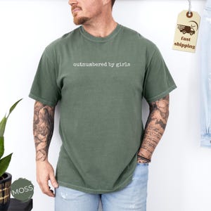 May include: A moss green t-shirt with the text "outnumbered by girls" in white. The short-sleeved shirt is made of a soft material. A fast shipping tag is visible in the upper right corner.