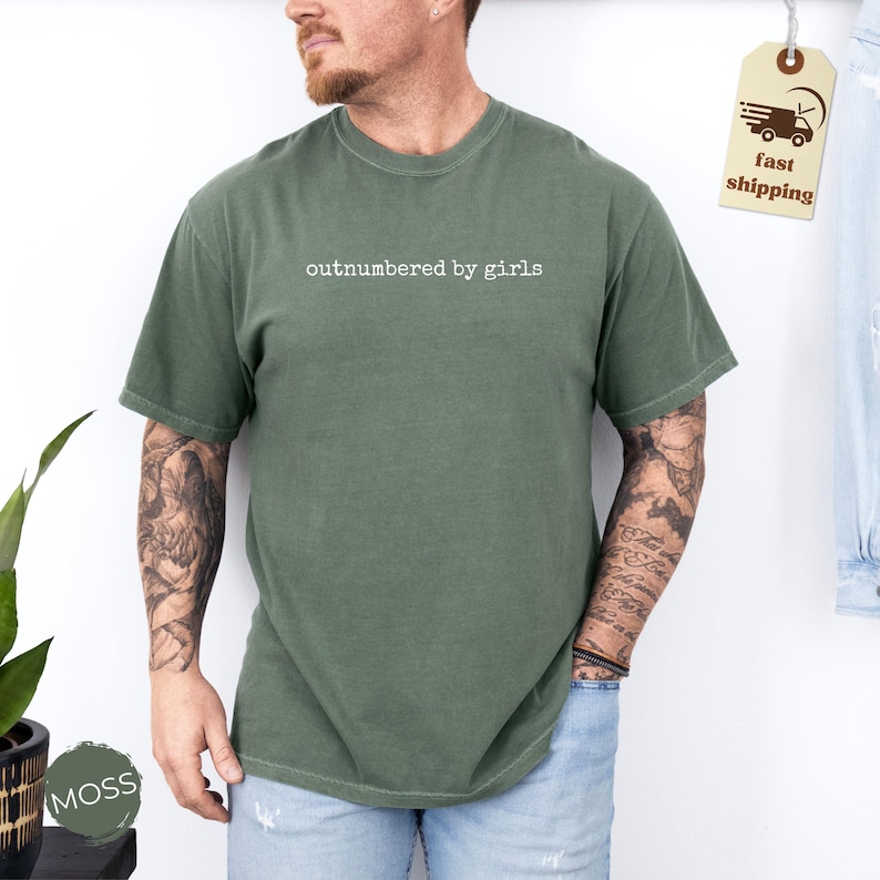 May include: A moss green t-shirt with the text "outnumbered by girls" in white. The short-sleeved shirt is made of a soft material. A fast shipping tag is visible in the upper right corner.