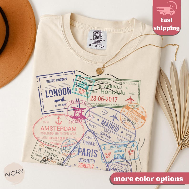 May include: Ivory-colored t-shirt with travel-themed stamp graphics in various colors. The shirt features stamps from London, Amsterdam, Paris, and other cities. A gold necklace and a hat are also visible.