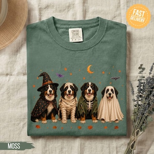 May include: A moss green t-shirt featuring a graphic of four Bernese Mountain dogs dressed in Halloween costumes: a witch, a mummy, a skeleton, and a ghost. The design includes a crescent moon, stars, and bats. The shirt has a Comfort Colors label.
