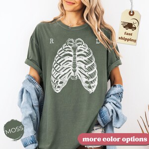 May include: Moss green t-shirt featuring a white rib cage graphic. The letters "R" and "L" are above the rib cage design. A tag with "fast shipping" is visible. The words "more color options" are at the bottom.