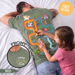 May include: Moss green t-shirt with a colorful fantasy map design on the back, featuring unicorns, a castle, and a rainbow path. The front of the shirt has the text "Tired Dads Club". A child is playing with toy figures on the shirt.