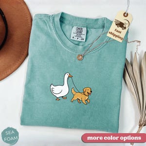 Pu&ograve; includere: T-shirt Comfort Colors verde acqua con una grafica di un'oca bianca e un golden retriever al guinzaglio. La maglietta ha un girocollo e una piccola etichetta con la scritta "fast shipping". Una collana d'oro e un cappello marrone sono anche nell'immagine.