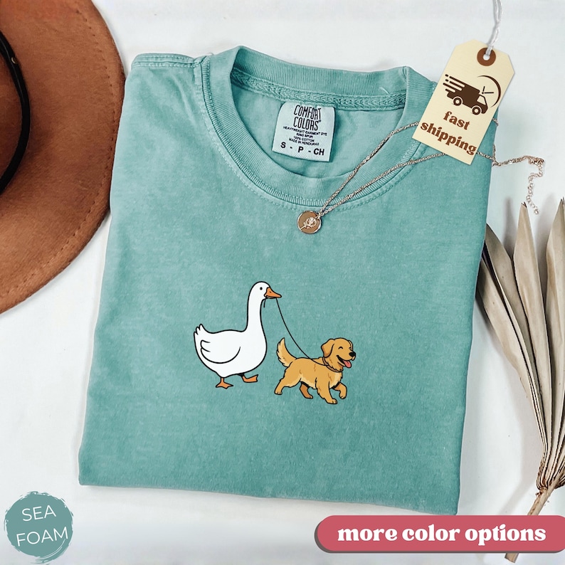 Pu&ograve; includere: T-shirt Comfort Colors verde acqua con una grafica di un'oca bianca e un golden retriever al guinzaglio. La maglietta ha un girocollo e una piccola etichetta con la scritta "fast shipping". Una collana d'oro e un cappello marrone sono anche nell'immagine.