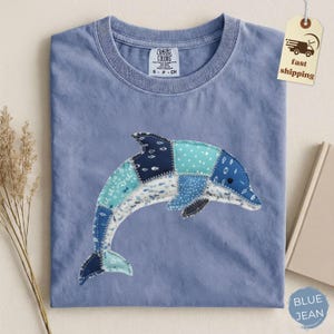 May include: A light blue Comfort Colors t-shirt featuring a patchwork dolphin design in various shades of blue and white. The shirt has a crew neck and short sleeves. A tag with "fast shipping" is attached.