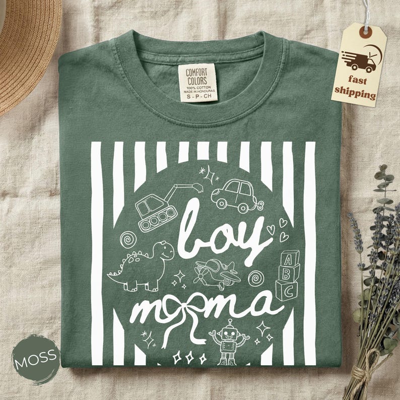 May include: A moss green t-shirt with a white graphic that says "boy momma" with illustrations of a dinosaur, car, airplane, robot, and more. The shirt is made of Comfort Colors cotton.