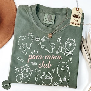 以下が含まれることがあります： モスグリーンのTシャツ。白いポメラニアン犬のグラフィックとピンク色の「pom mom club」の文字がプリントされています。Tシャツはクルーネックで半袖です。ペンダント付きのゴールドネックレスがTシャツの上にあります。