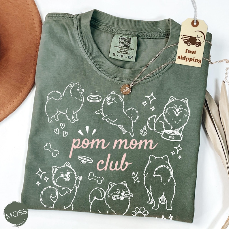 以下が含まれることがあります： モスグリーンのTシャツ。白いポメラニアン犬のグラフィックとピンク色の「pom mom club」の文字がプリントされています。Tシャツはクルーネックで半袖です。ペンダント付きのゴールドネックレスがTシャツの上にあります。