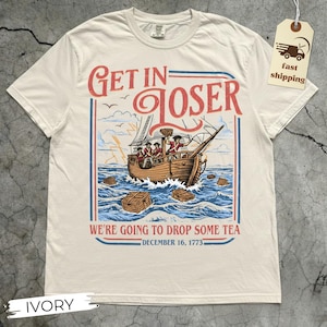 May include: Ivory-colored t-shirt with a vintage-style graphic. The design features a ship on the water with the text "GET IN LOSER" above and "WE'RE GOING TO DROP SOME TEA DECEMBER 16, 1773" below. A tag with "fast shipping" is attached.