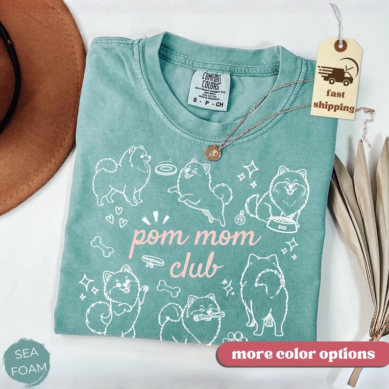 以下が含まれることがあります： ポメラニアン犬の白いグラフィックと「pom mom club」の文字が入った、シーフォームグリーンのTシャツ。 Tシャツはクルーネックと半袖です。 金色のネックレスがシャツの上にかけられています。