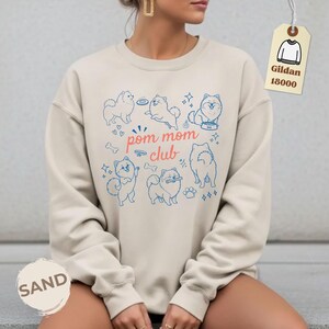 以下が含まれることがあります： サンドカラーのスウェットシャツ。青いポメラニアン犬のイラストと「pom mom club」の文字が描かれています。デザインにはハート、骨、星が含まれています。Gildan 18000のタグが付いています。