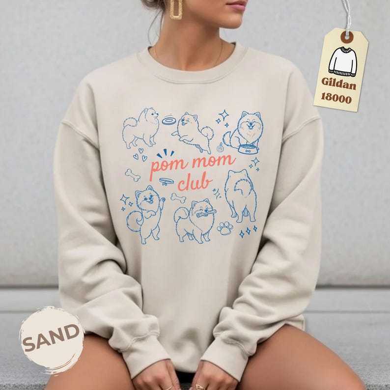 以下が含まれることがあります： サンドカラーのスウェットシャツ。青いポメラニアン犬のイラストと「pom mom club」の文字が描かれています。デザインにはハート、骨、星が含まれています。Gildan 18000のタグが付いています。
