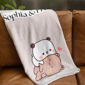 May include: A soft, light pink throw blanket with a white border. The blanket features a cute cartoon panda bear hugging a brown bear. The text "Sophia &" is printed on the top of the blanket.